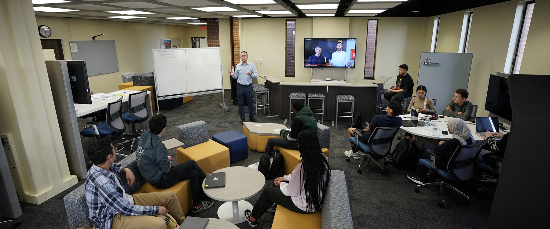 A group of students sit on couches an chairs in a large room filled with whiteboards, TVs and desks, listening to their professor lecture. 