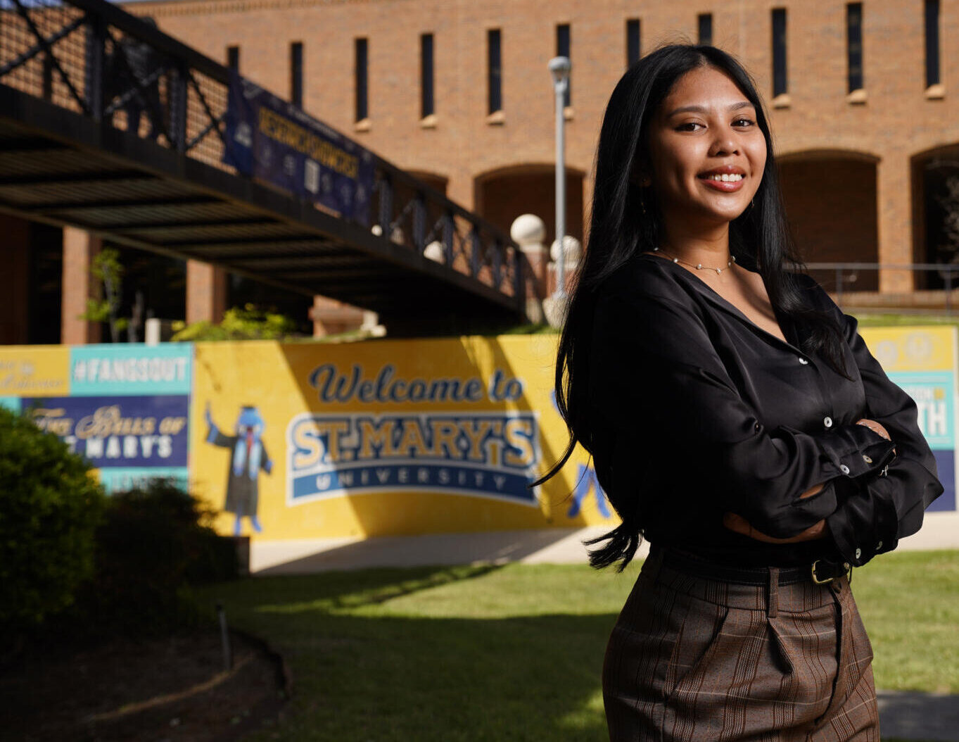 Female student smiles as she stands in front of the St. Mary's University Spirit wall. On the wall, the words "Welcome to St. Mary's University" can be seen with the school mascot, Rattler Man.