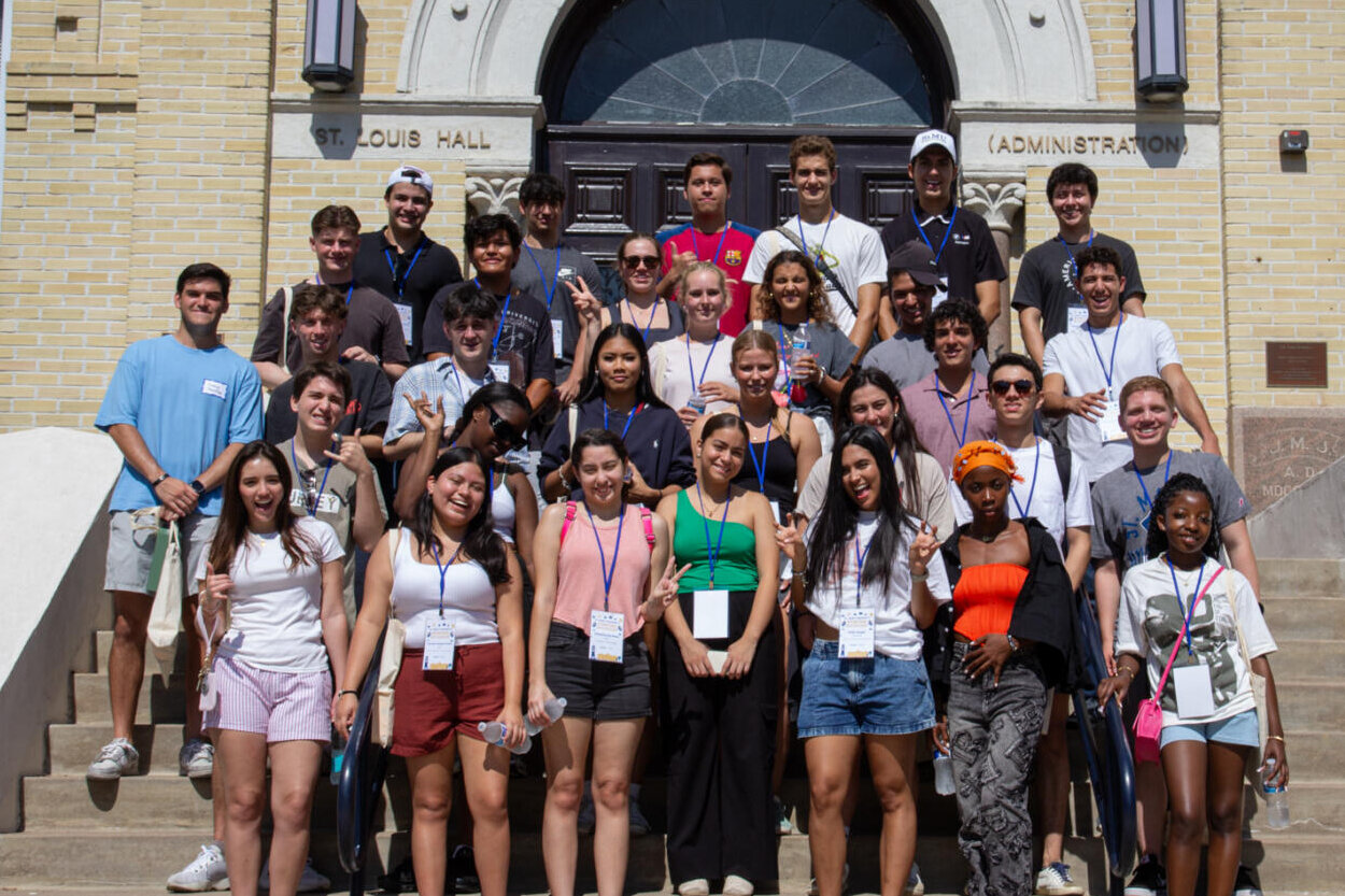 Students smile and wave their hands as they take a photo in front of St. Louis Hall at St. Mary's University.
